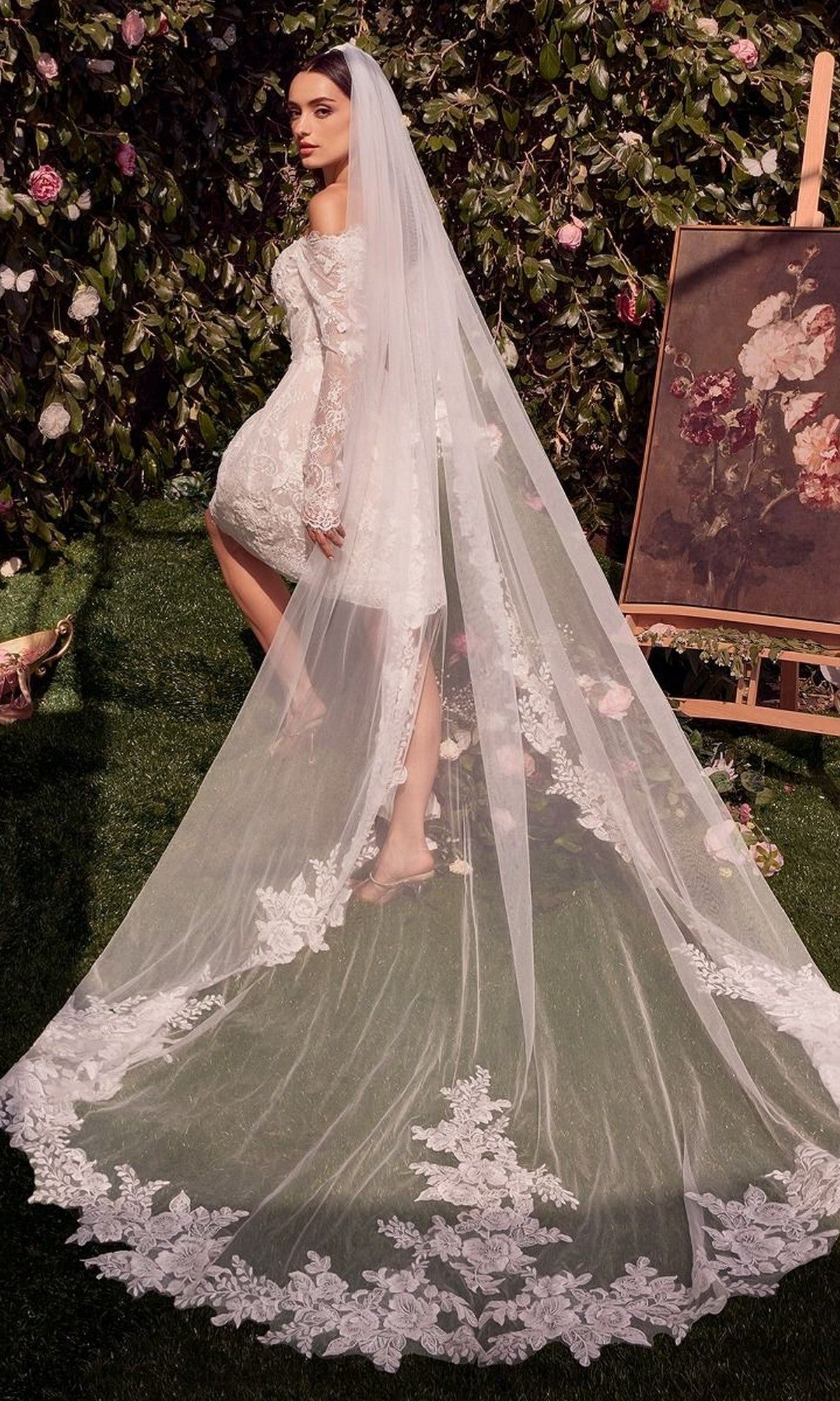 Wearing the Andrea and Leo WL021 short white dress with an off-the-shoulder sweetheart neckline and fitted corset bodice, a woman stands outdoors near an easel and floral painting, her embroidered veil trailing behind.